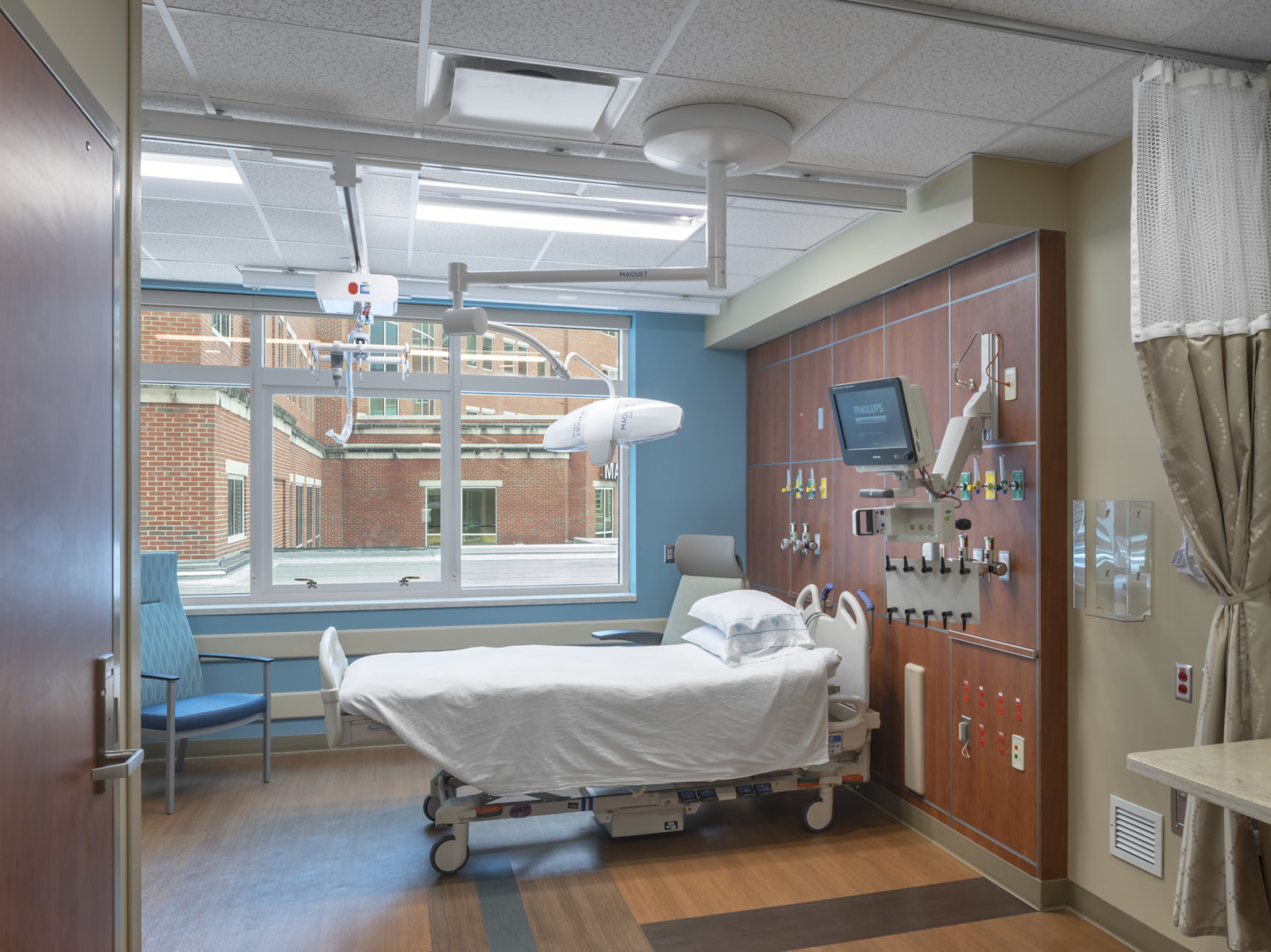 Hospital room with wood flooring and accent wall panel over bed. Large window looks out to exterior and brick wall