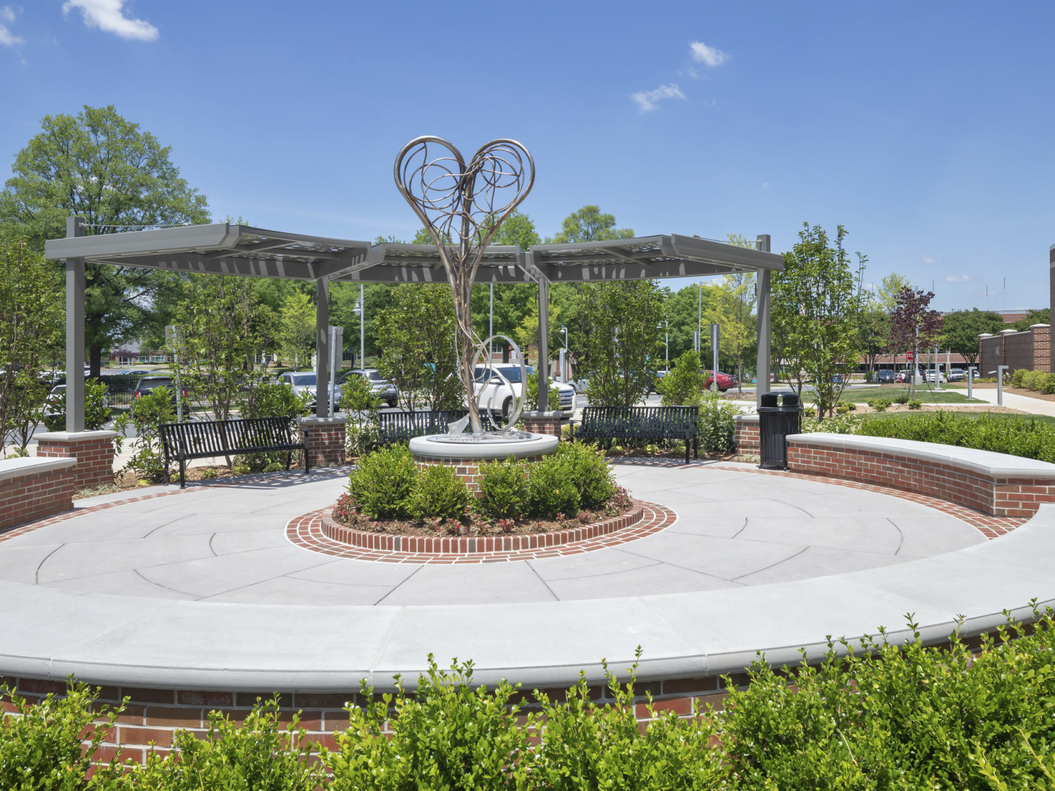 Exterior paved round seating area in garden lined with brick bench wall and covered benches. Abstract heart shaped sculpture, center