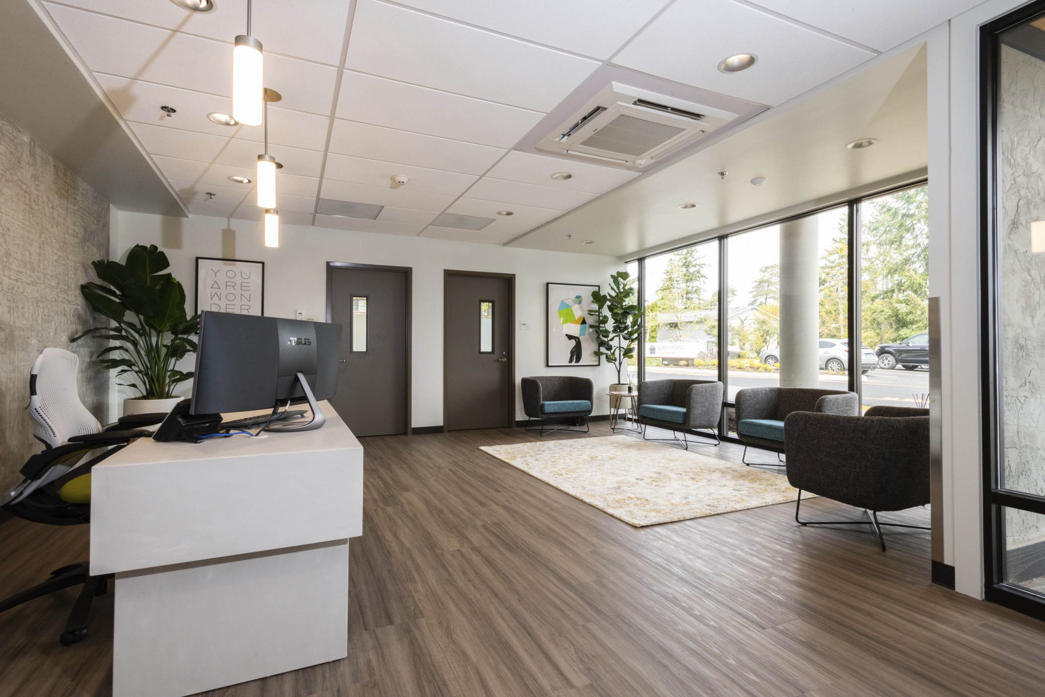 reception room with white desk under pendant light, wood floors, floor to ceiling glass. beige area rug anchors soft seating