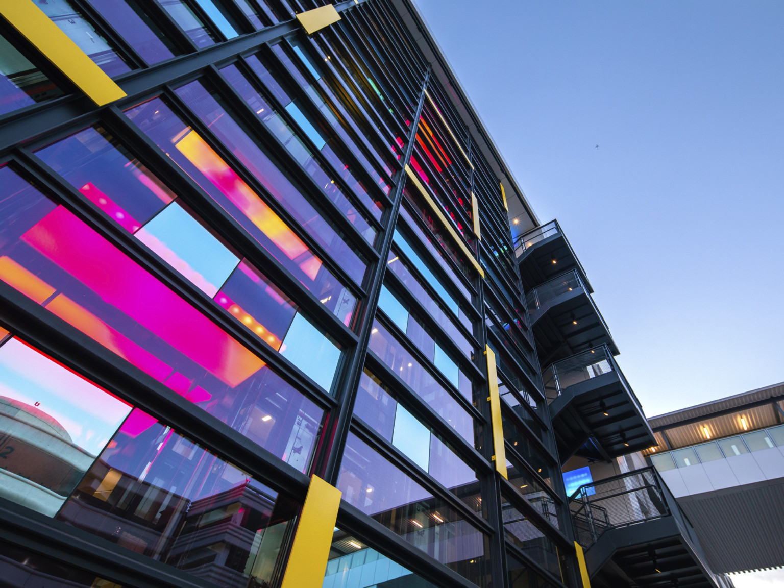 View looking up at central glass tower with iridescent window panels and staircase to the right. Skywalk over road to right