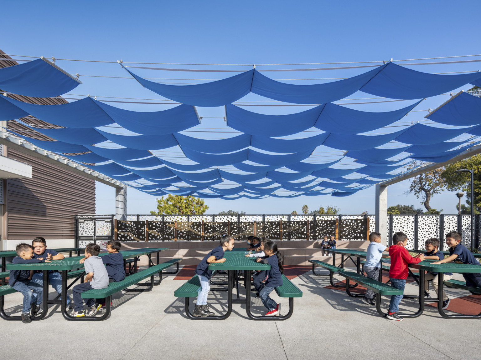 Outdoor seating area shaded from above by draping blue panels of fabric. Students sit at rows of green picnic tables, white fence