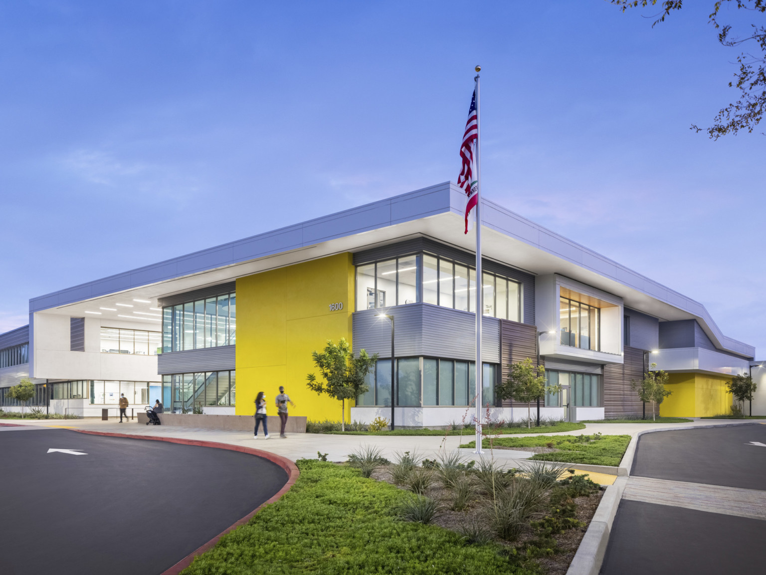 Corner view of yellow and grey 2 story school with large windows, landscaping and american flag, illuminated in the evening