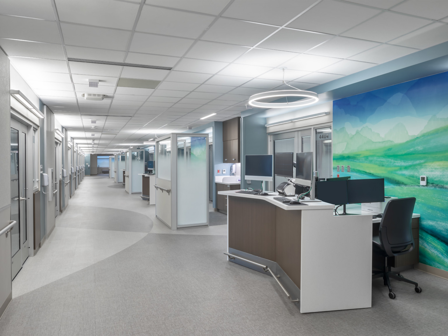 Hospital corridor with angled desk with multiple monitors in front of abstract blue and green mural. Glass partitions beyond