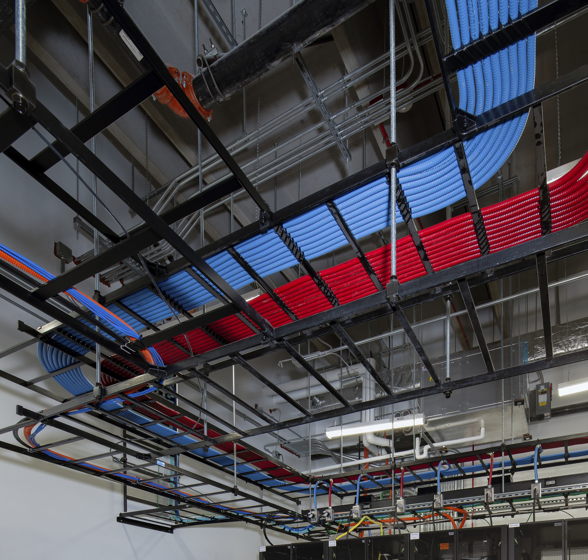 an open ceiling with black orthogonal framing system that contains lighting pendants, pipes, metallic and red and blue tubes