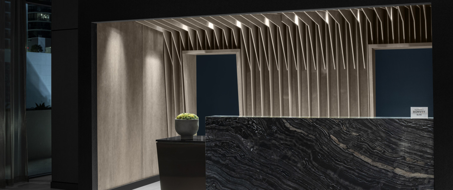 The AC Hotel reception desk, a dark stone slab with warm wood slats like palm fronds vertically emerge overhead in recessed space