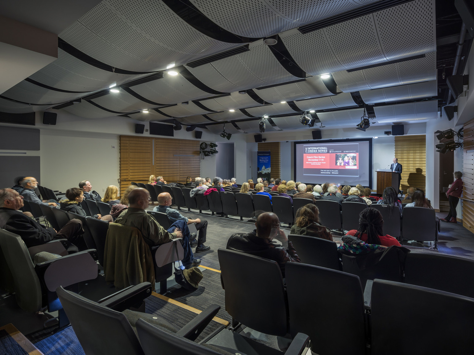 lecture room with curved perforated acoustic ceiling panels projection screen and wood panels on walls gray carpet and seats