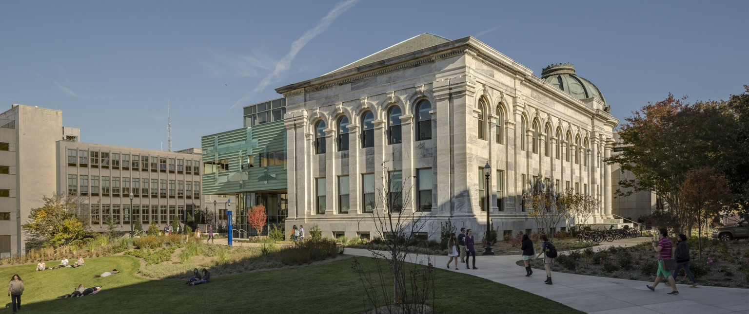 neoclassical stone building with arched windows, columns, radial stairs. modern glass and teal green facade cladding addition