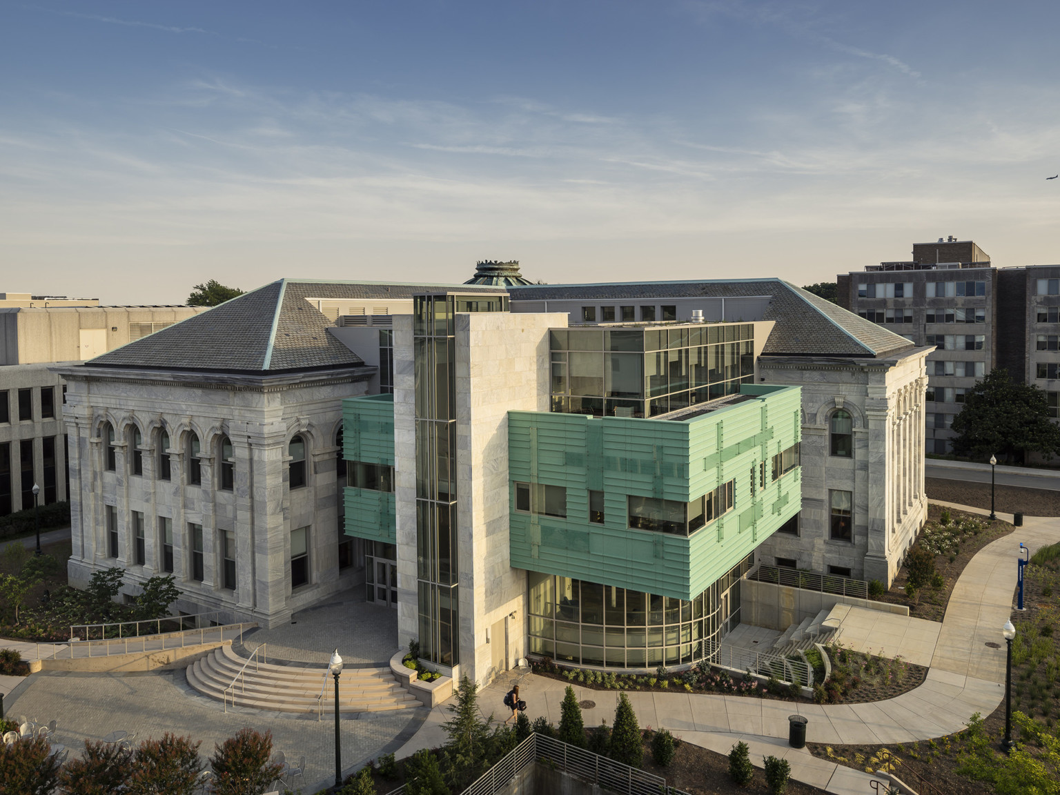 L shaped neoclassical gray and white striated marble building with modern glass and green facade clad volume in the middle