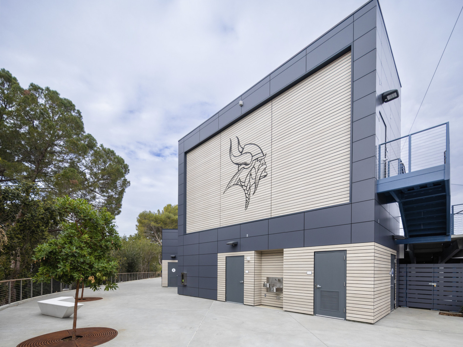 Back view of press box with Viking logo embossed into phenolic panels. Grey paneled wrap facade. Courtyard with bench and tree