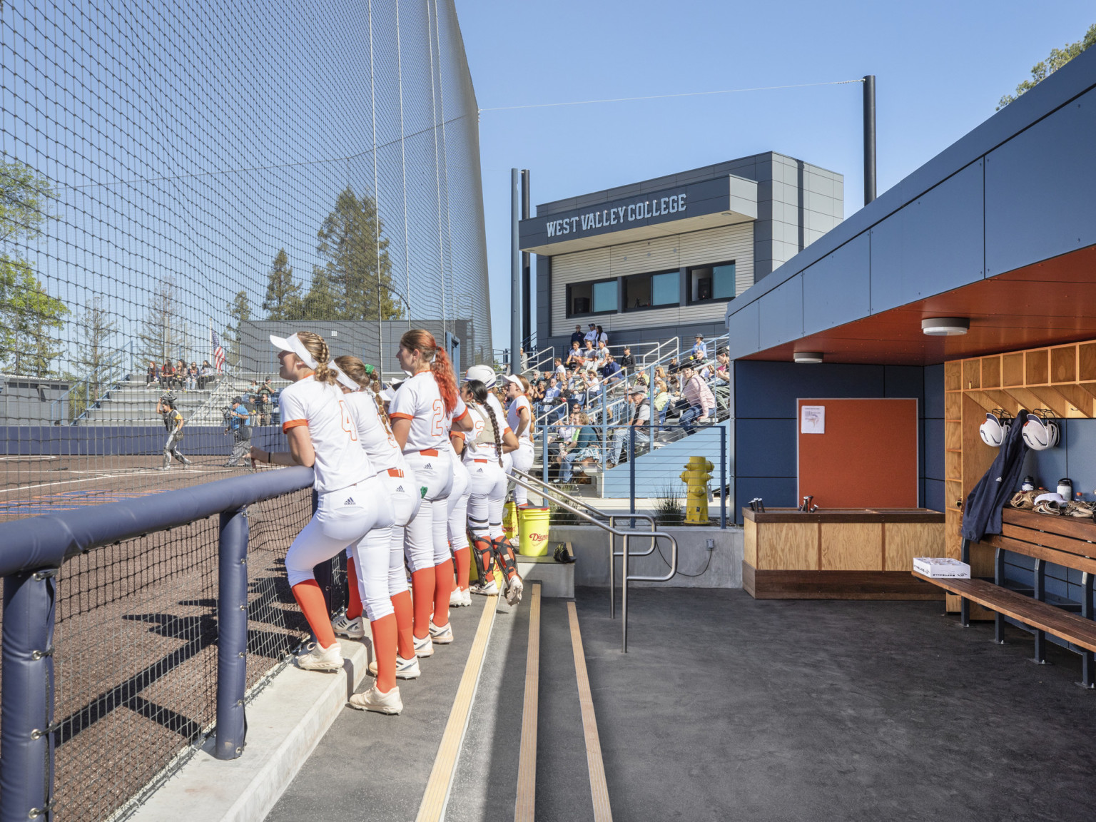 A row of girls stand at the railing along dugout watching softball game. Grey panel wrap facade with wood shelving and cabinets