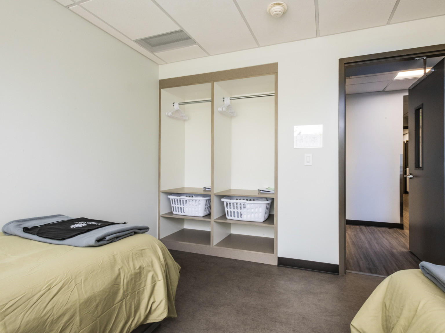 a room with white walls and closets with hangers, laundry baskets, and shelves. A wooden door leads to a wood floor hallway