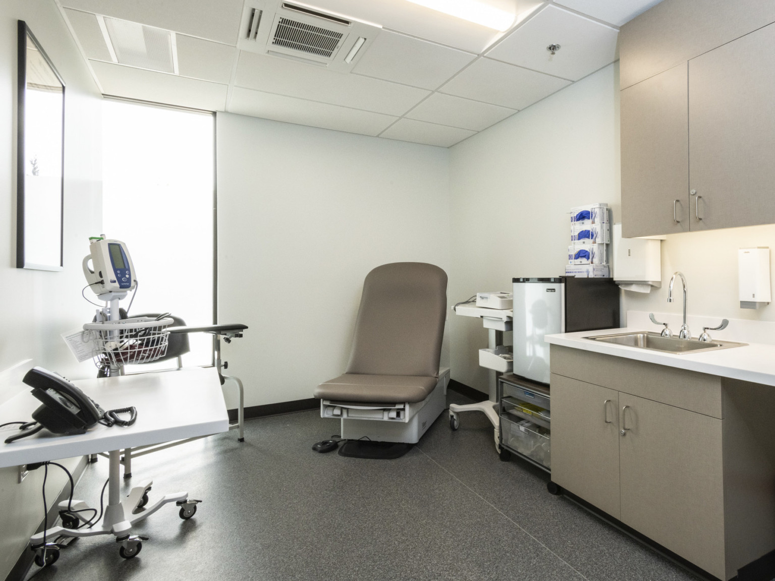 medical exam room with medical furniture on casters white ceiling and walls gray floor and tan cabinet storage and exam chair