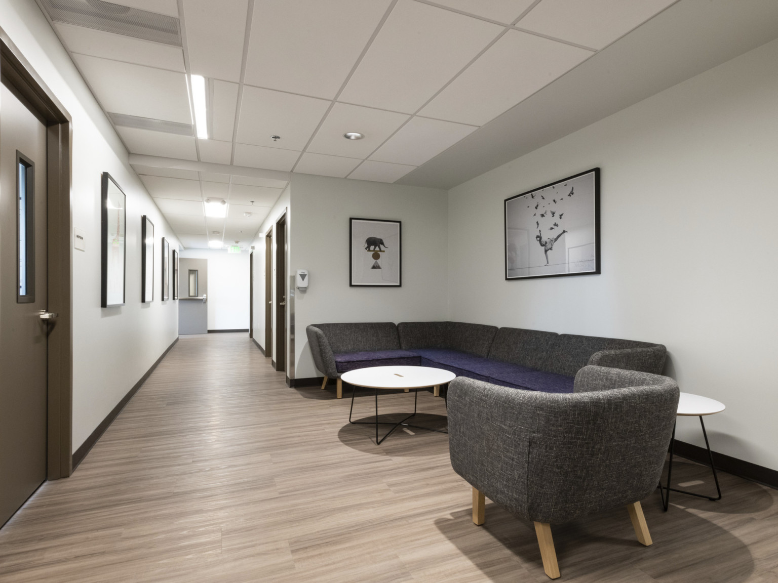 seating area with gray and purple sofa and tables in a hallway lined with photo and brown doors with rectangular windows