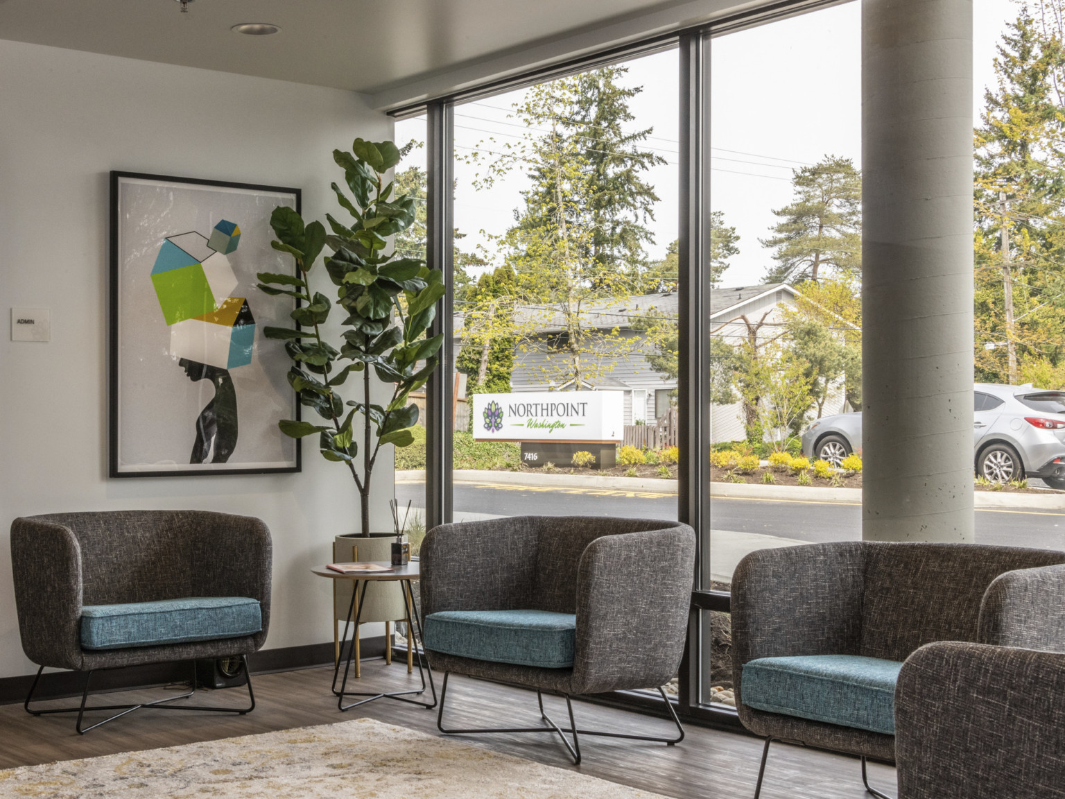 a seating area with gray upholstered bucket seating and blue seat cushions in front of floor to ceiling glass & abstract art