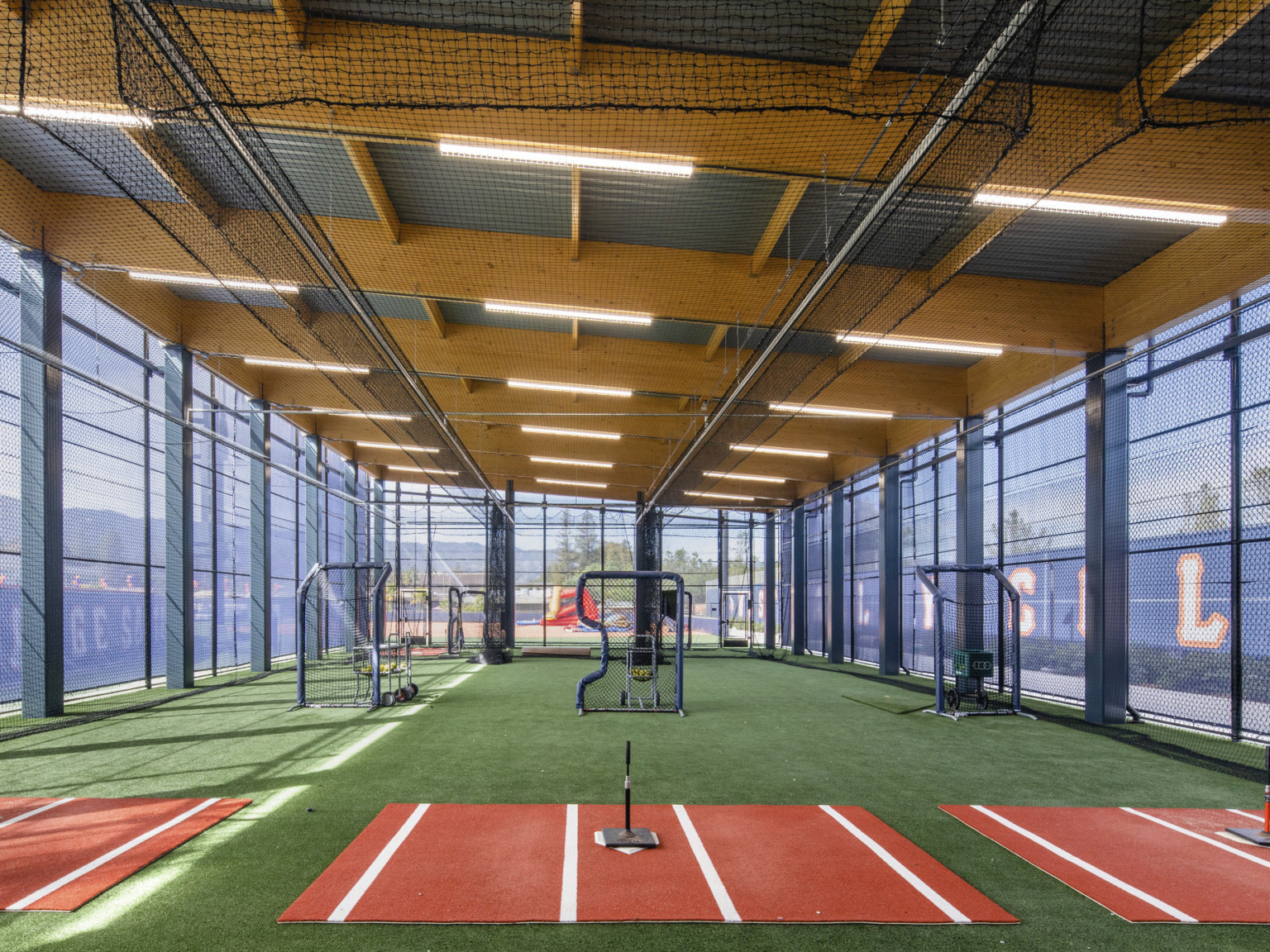 Training space with exposed wood beams overhead. Turf ground with batting area and bins of balls behind netting