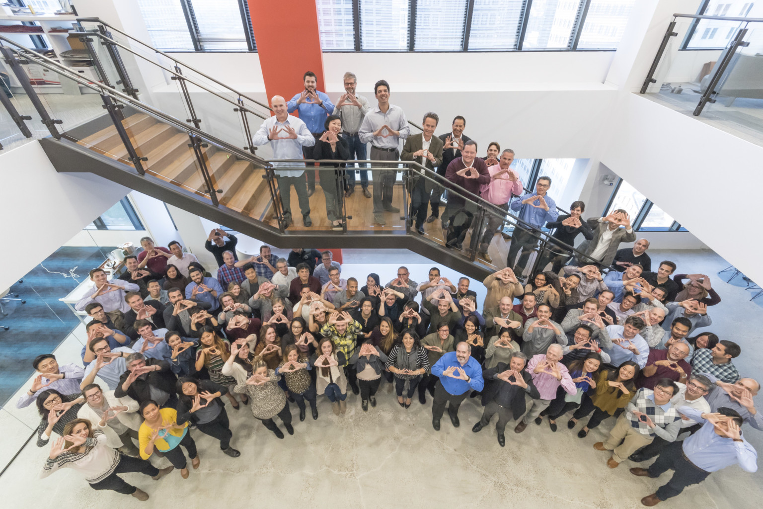 large group of professionals standing in double height space on a beige floor and ascending a wood stair with glass rails