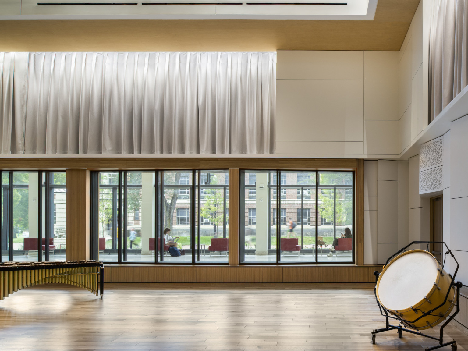 Double height practice room of music school with instruments. Large interior windows look to hallway and courtyard beyond