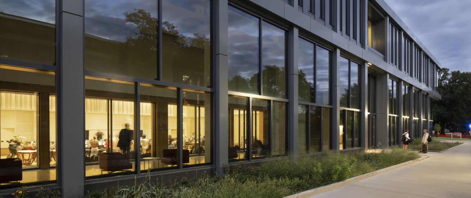 Exterior of Timashev Family Music Building viewed from plant lined sidewalk with view into music practice room illuminated within
