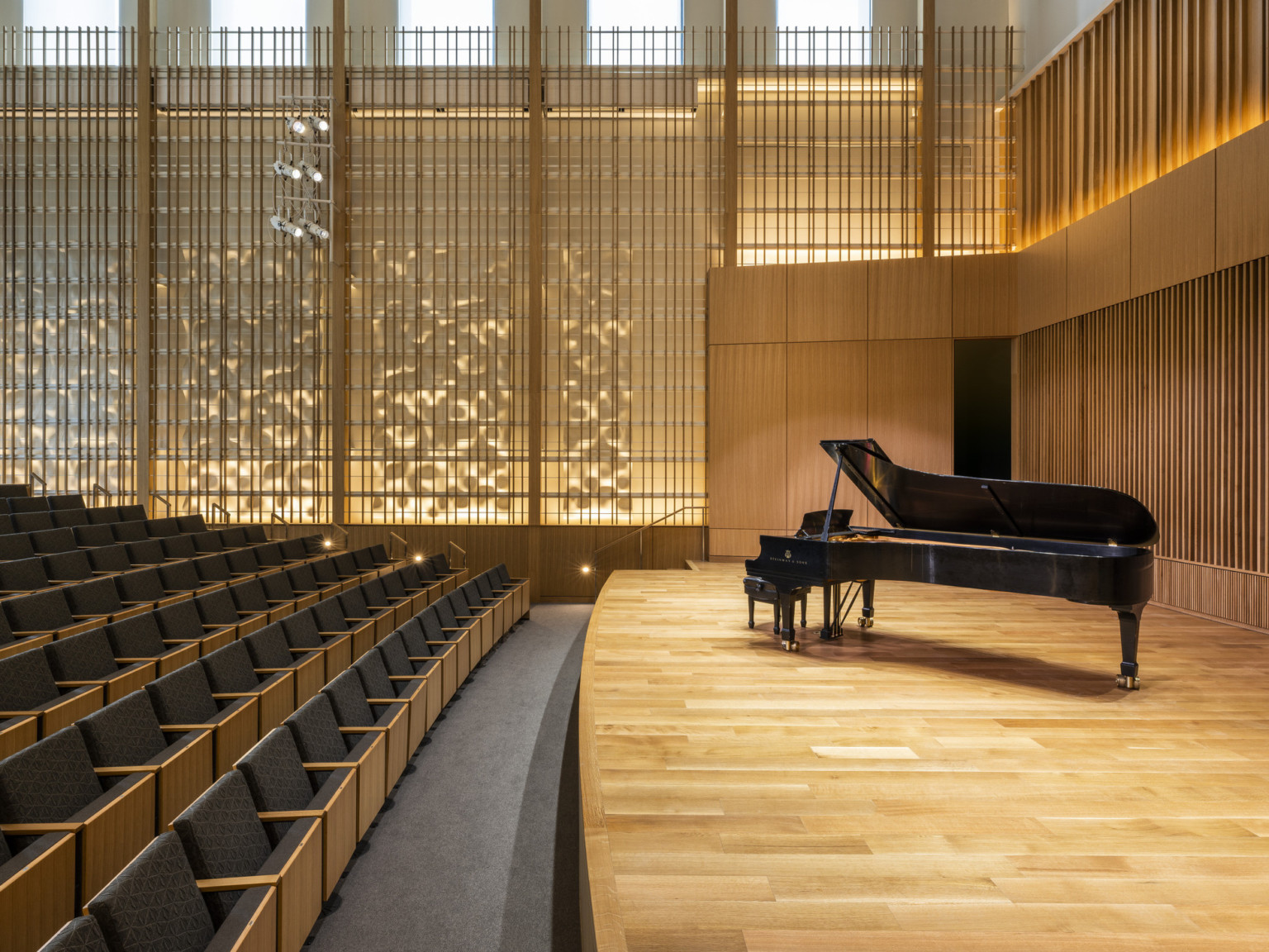Angled view from side of stage looking toward wood design on wall illuminated from behind. Grey theater seats with light wood