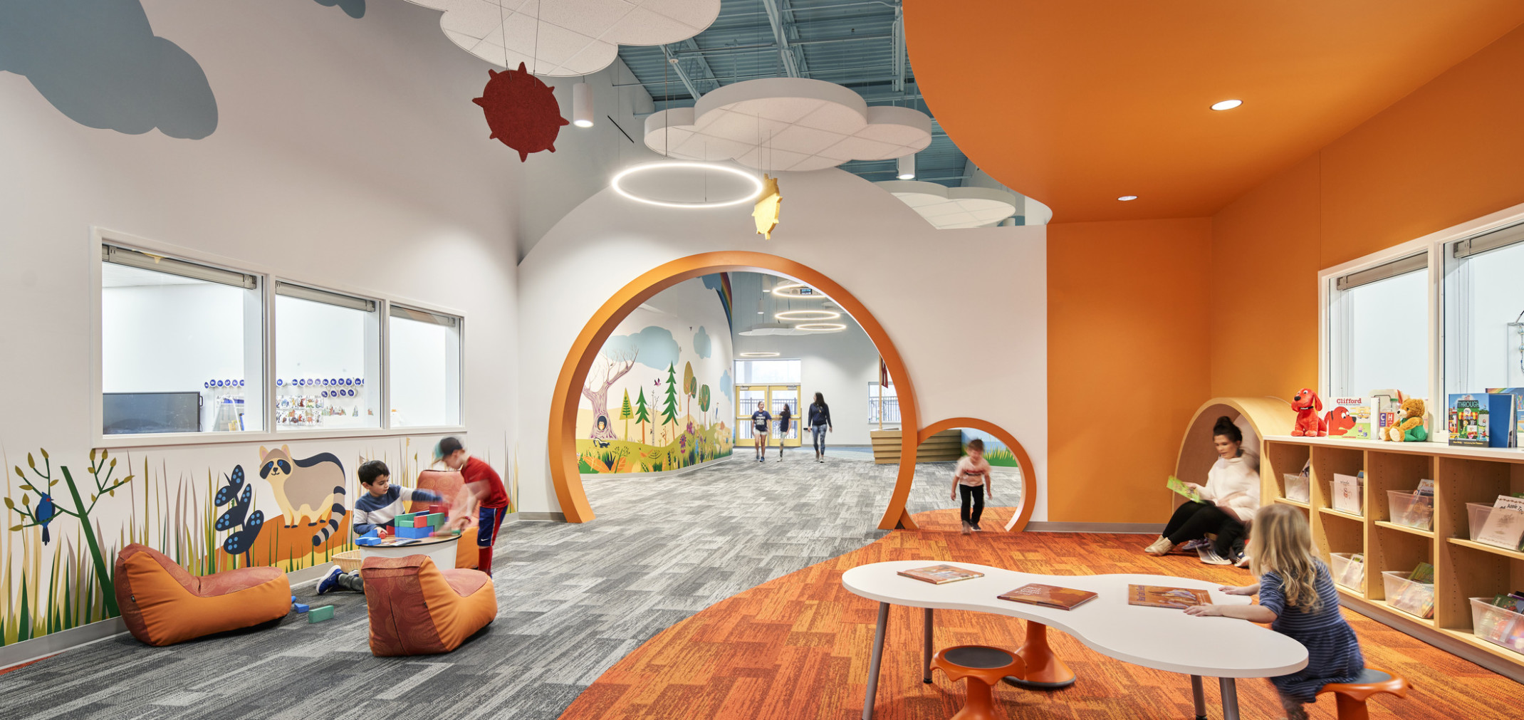 A vibrant children's library with orange and grey décor, featuring playful murals, hanging decorations, and children engaging in activities.