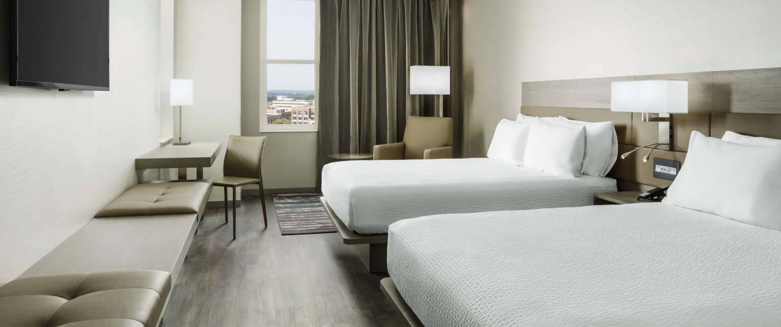 Guest room with two white beds divided by an end table and light fixture. Arm chair by window, all face bench along left wall