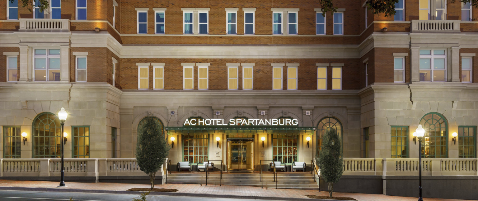 Exterior of hotel entry with sign illuminated on green awning. Green arches windows on carved front stone wall