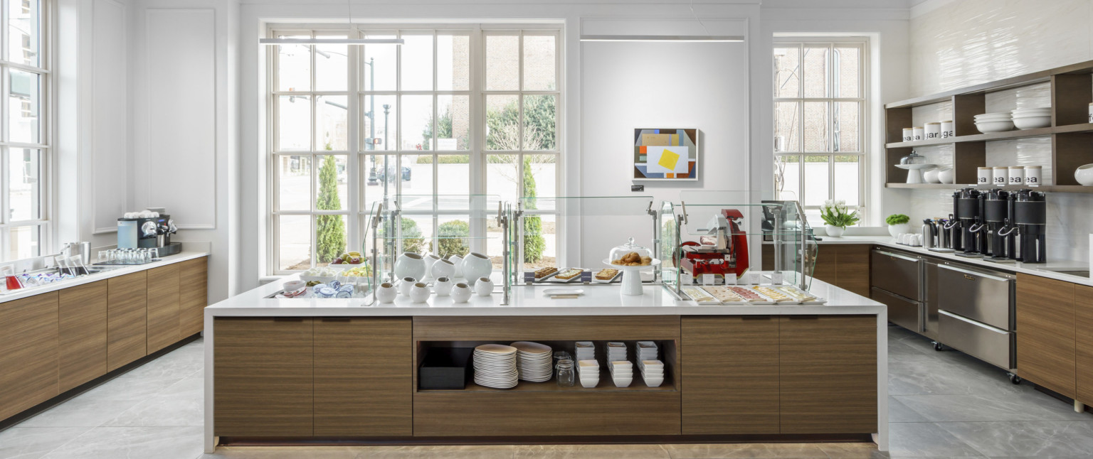 White counter with wood storage underneath set for a breakfast buffet in a white room with large windows on ground level