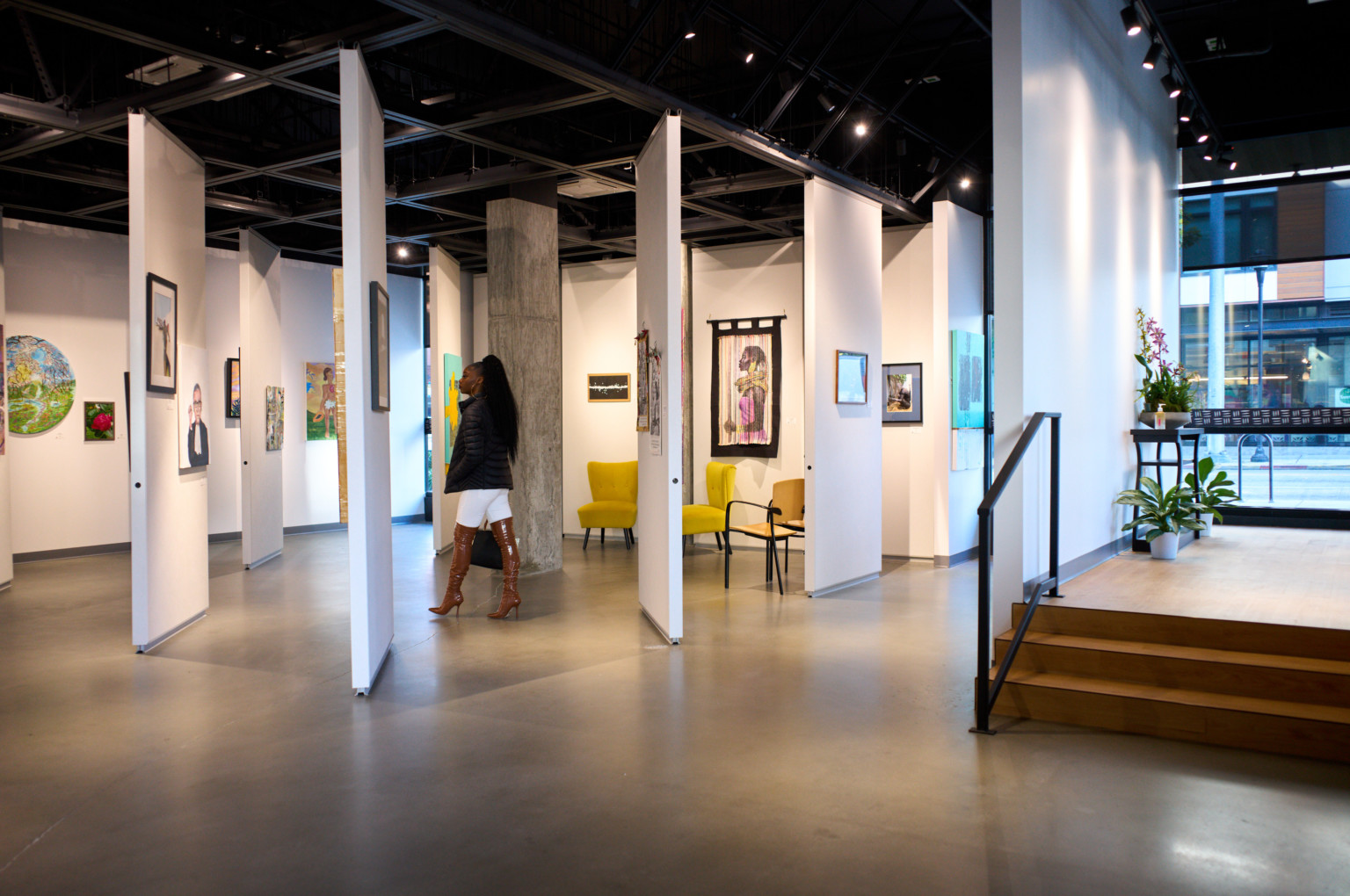 Woman standing in Arte Noir Gallery interior in Seattle, movable white wall panels hang art in room with floor to ceiling windows, black ceiling