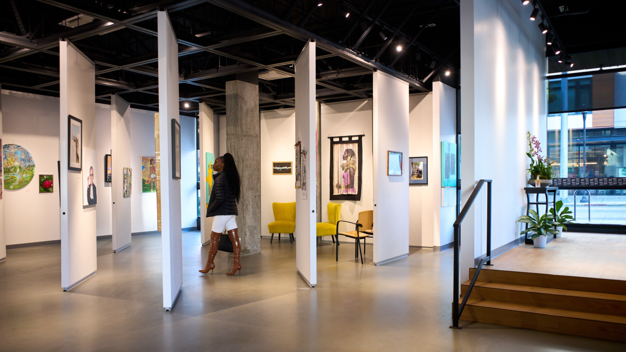 Woman standing in Arte Noir Gallery interior in Seattle, movable white wall panels hang art in room with floor to ceiling windows, black ceiling in an art gallery