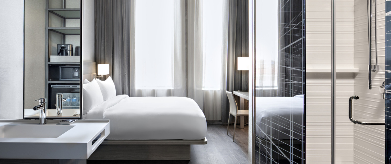 Guest room with white bed partially separated from kitchenette by translucent and metal wall right. Sink with mirror left