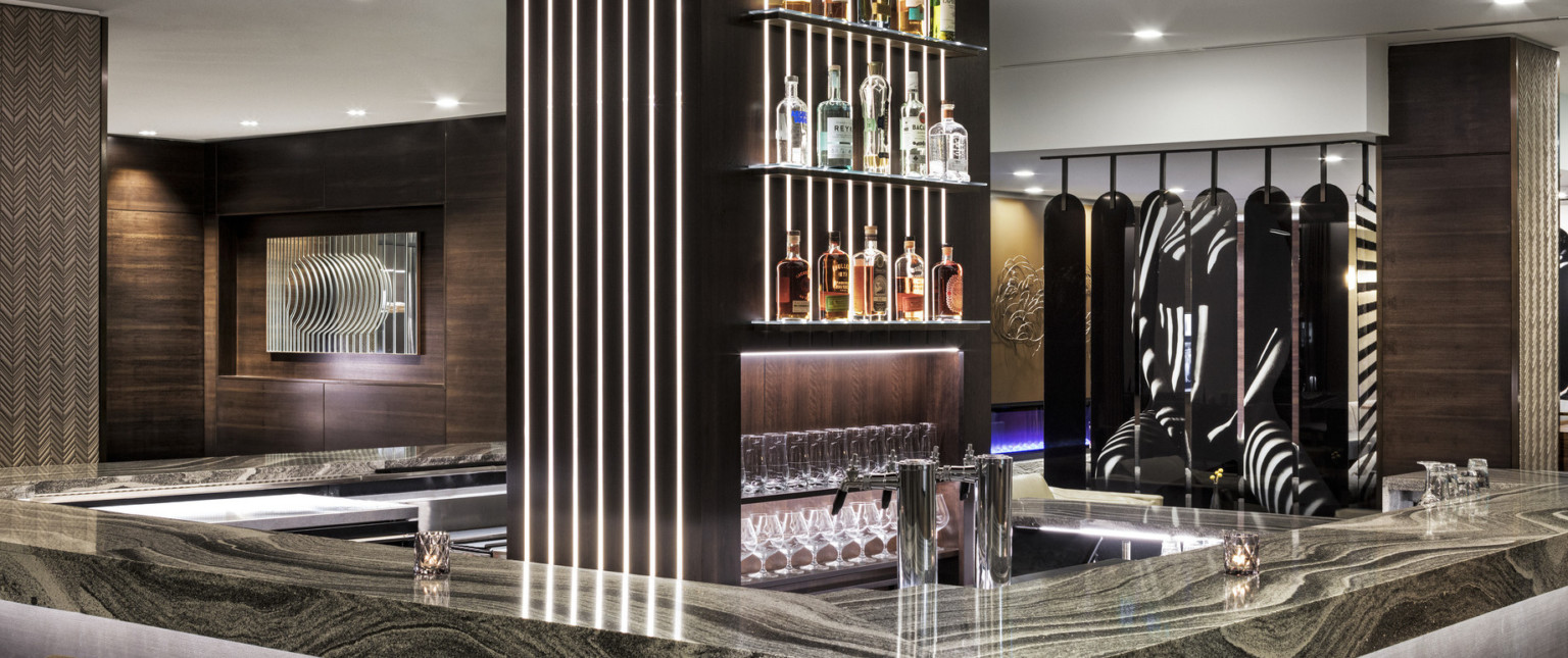 Rounded bar with dark marble counter and central wood pillar with shelving and illuminated recessed strips in wood panel room