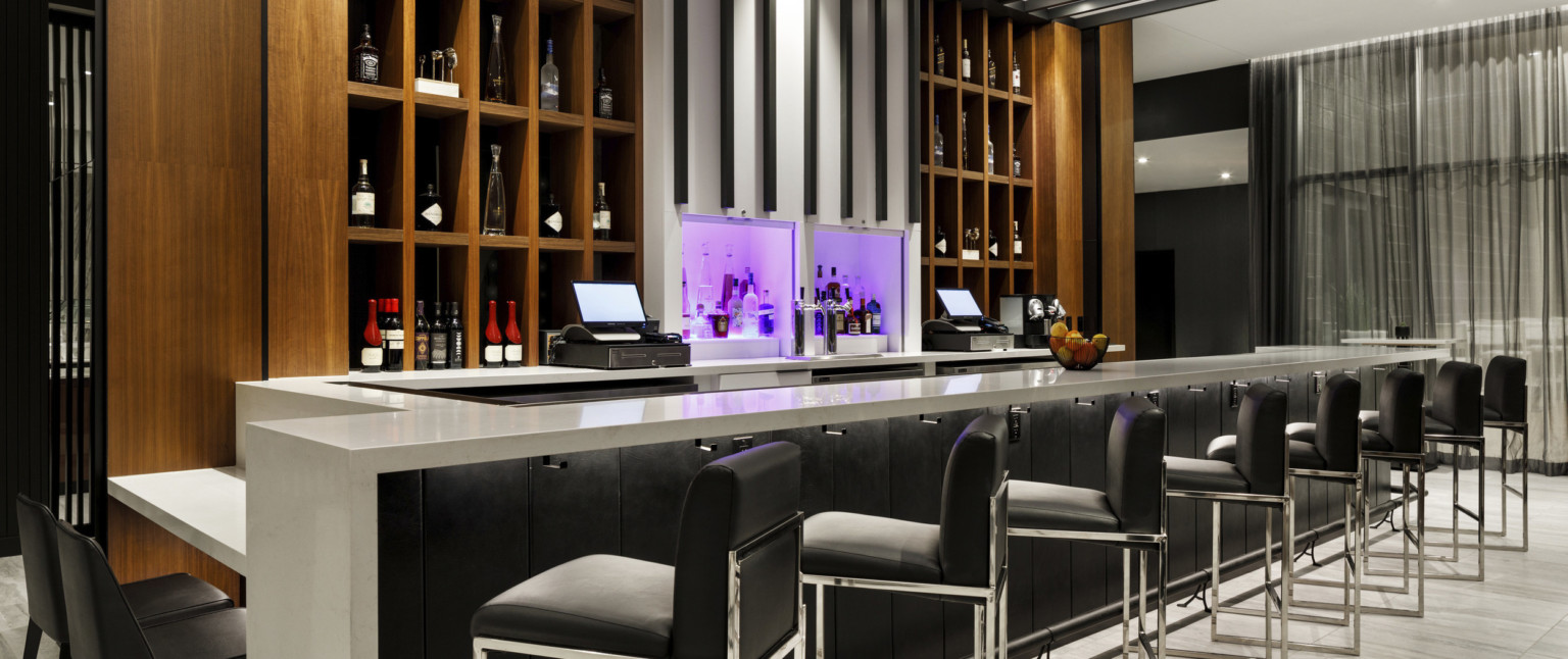 Bar with grey counter, black panel base, black bar chairs. Built in shelves behind bar, and white center column lit in purple