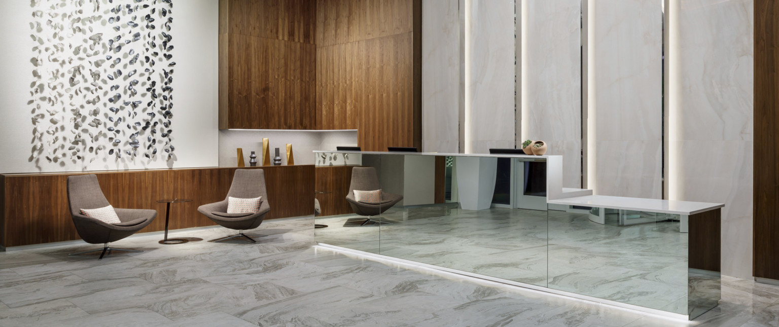 Mirrored reception desk with white counter in white lobby. Wood counter on left wraps up the wall at the corner near seating