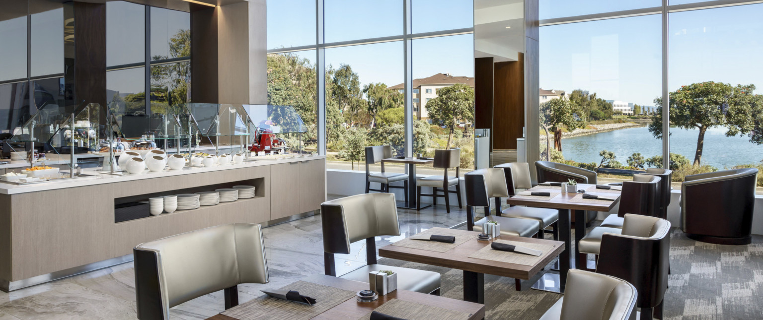 Seating area with set tables and double height windows. White counter with wood storage underneath is set for breakfast