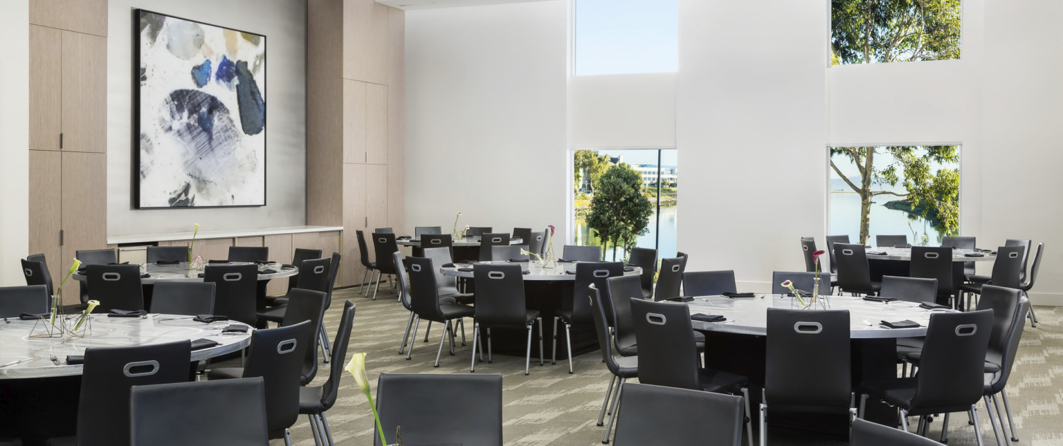 Carpeted seating area with round tables and black chairs. Wood cupboards on left wall with white counter space and framed art
