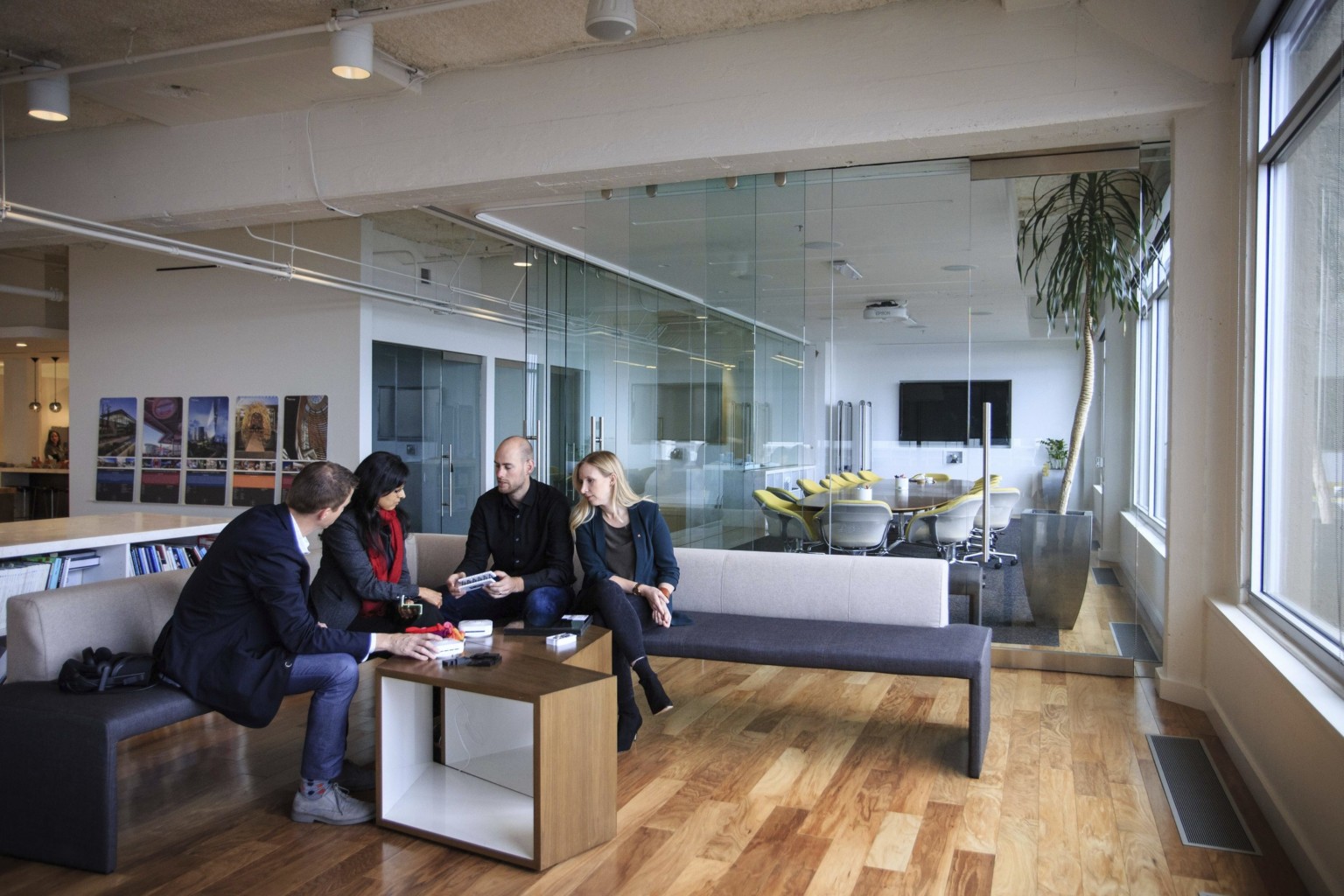 four professionals meeting over low angular wooden tables in a room with hardwood floors, broad windows, white walls