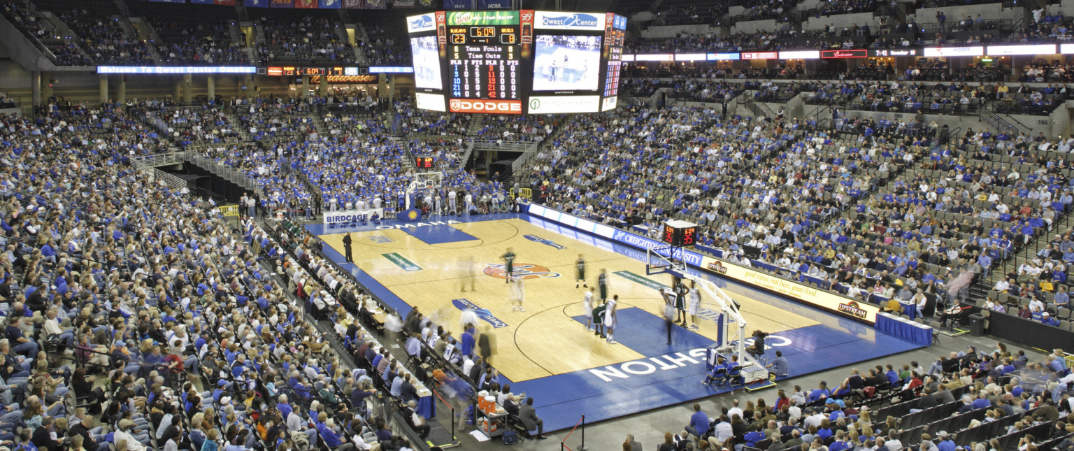 Basketball court with lighting and jumbotron hanging above. Bleachers surround the court and are filled with fans