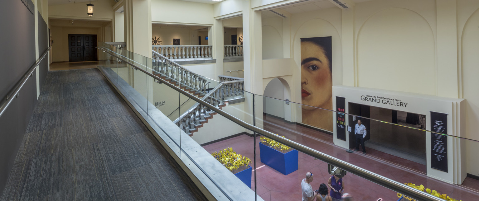 A carpeted walkway overlooks a staircase and an entryway labeled Grand Gallery in cream colored room with skylights