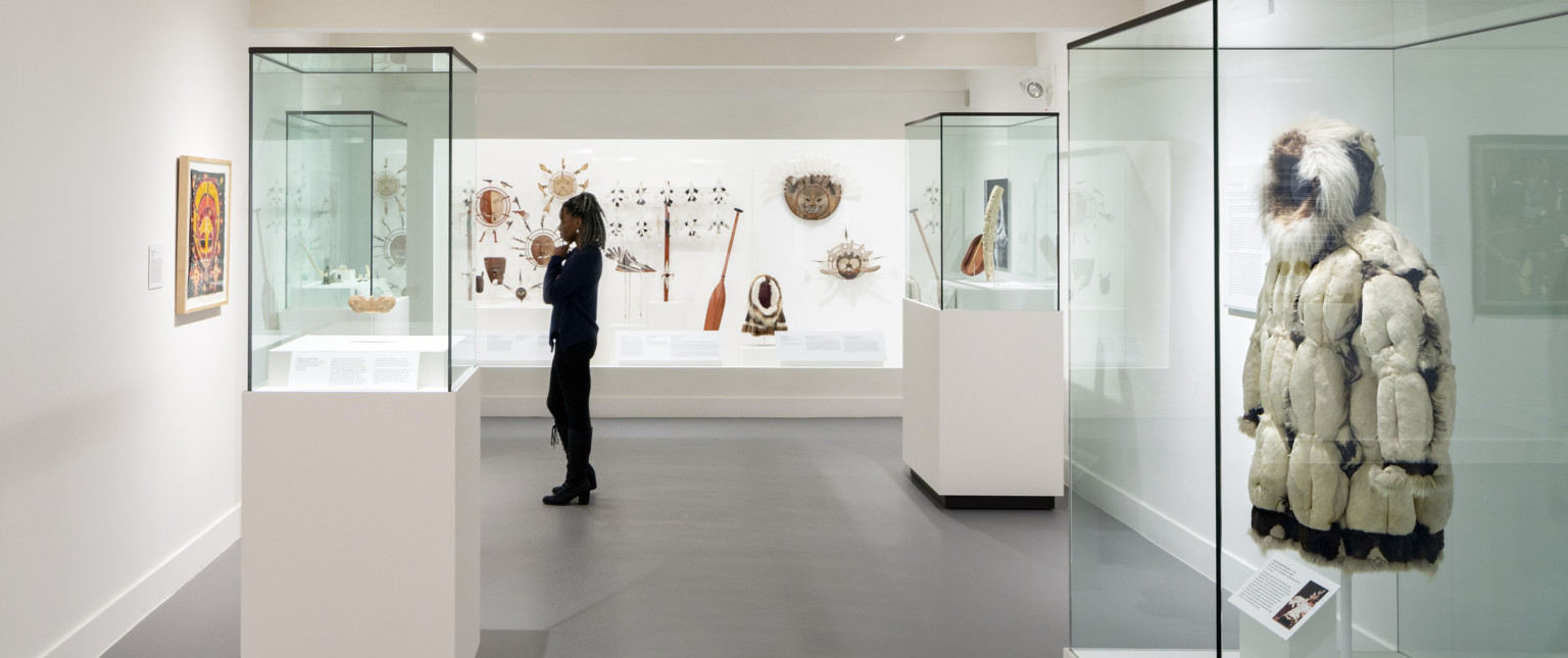 A gallery with traditional Native American clothing and artifacts, under a wood ceiling with exposed black support beams and pipes