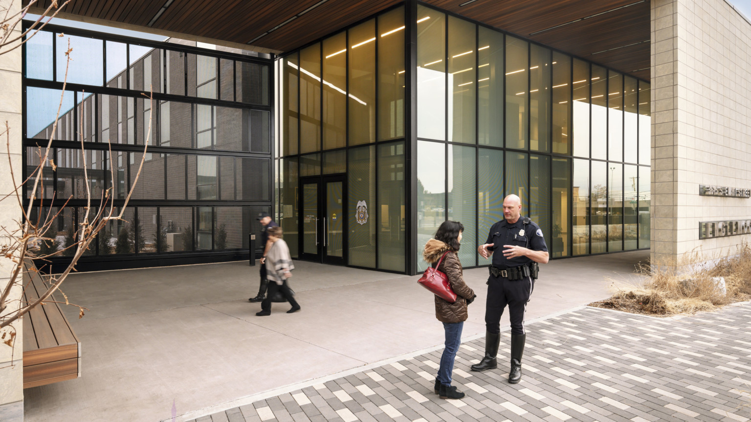 Police officer speaks with woman outside double height glass canopied entrance to Englewood Police Headquarters