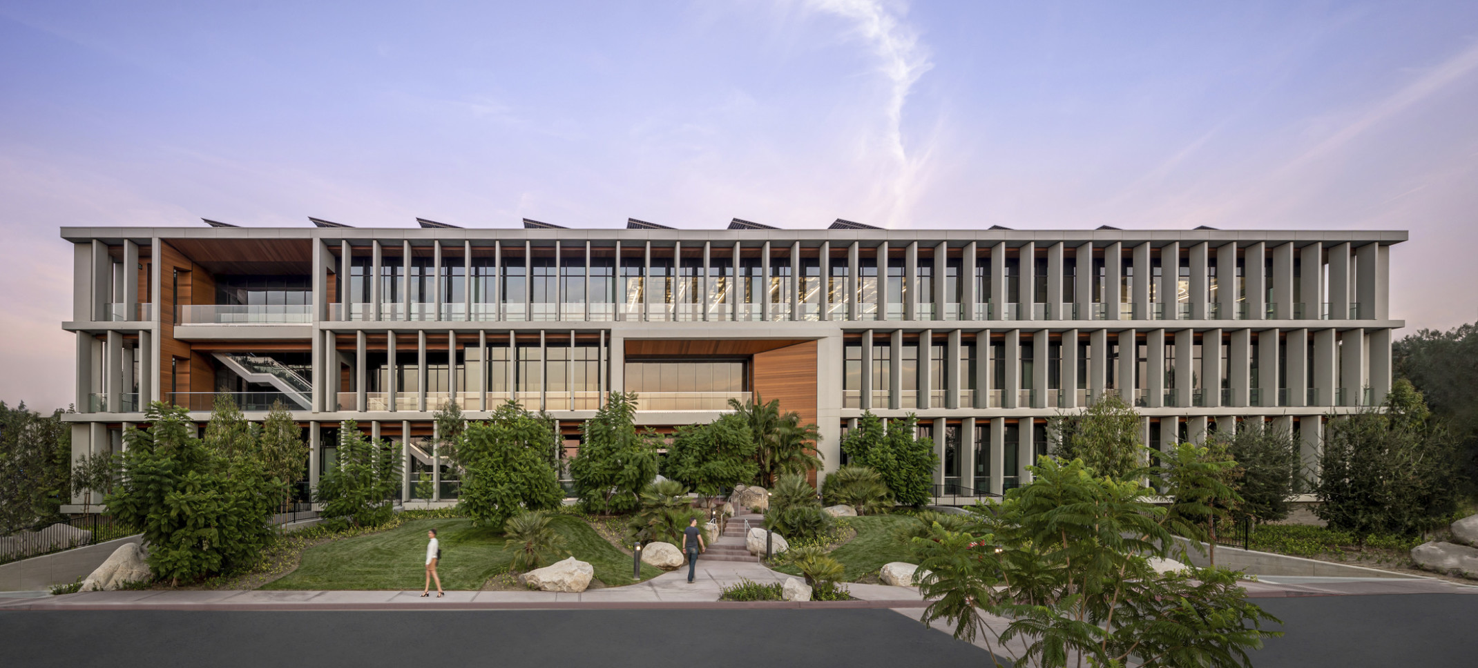 White wrapped slat facade with wood accents over above grade glass building. Lawn with large plants slopes up to building