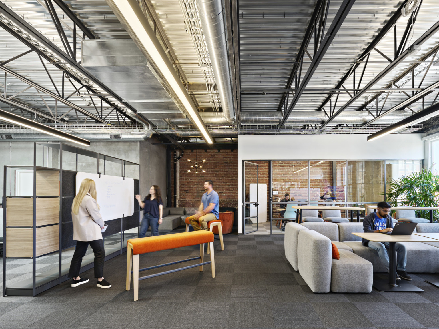 Office common space with mixed seating, benches and chairs. Flexible shelf wall with attached white board. Exposed brick, industrial ceiling