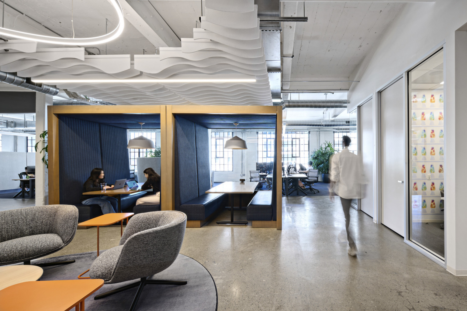 Office common space with grey armchairs and orange tables. 2 wood booths with bench seating at center enclosed front, back, above