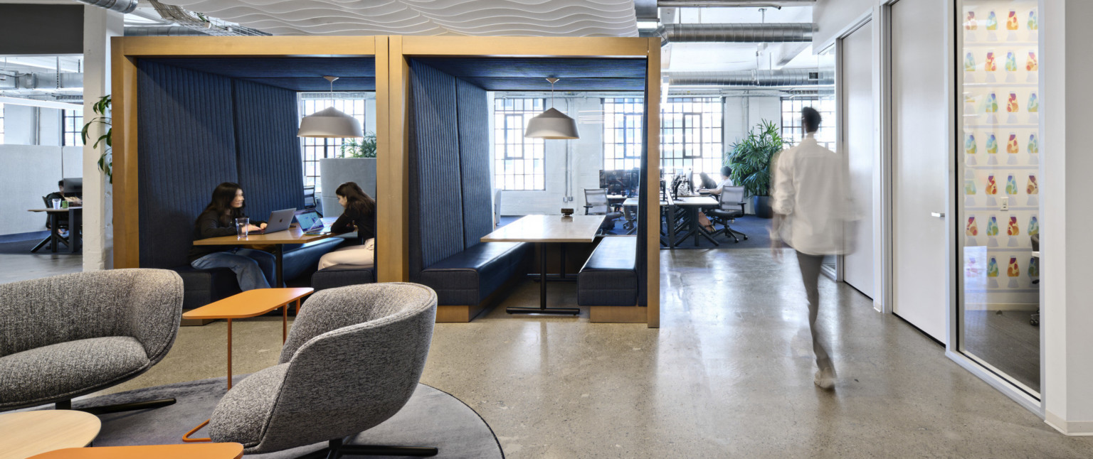 Office common space with grey armchairs and orange tables. 2 wood booths with bench seating at center enclosed front, back, above