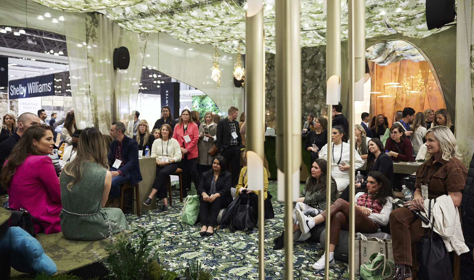 Inside the booth, green floral drapes hang over mixed seating. Crowd faces speakers, left. sculptural lighting. Orange next section