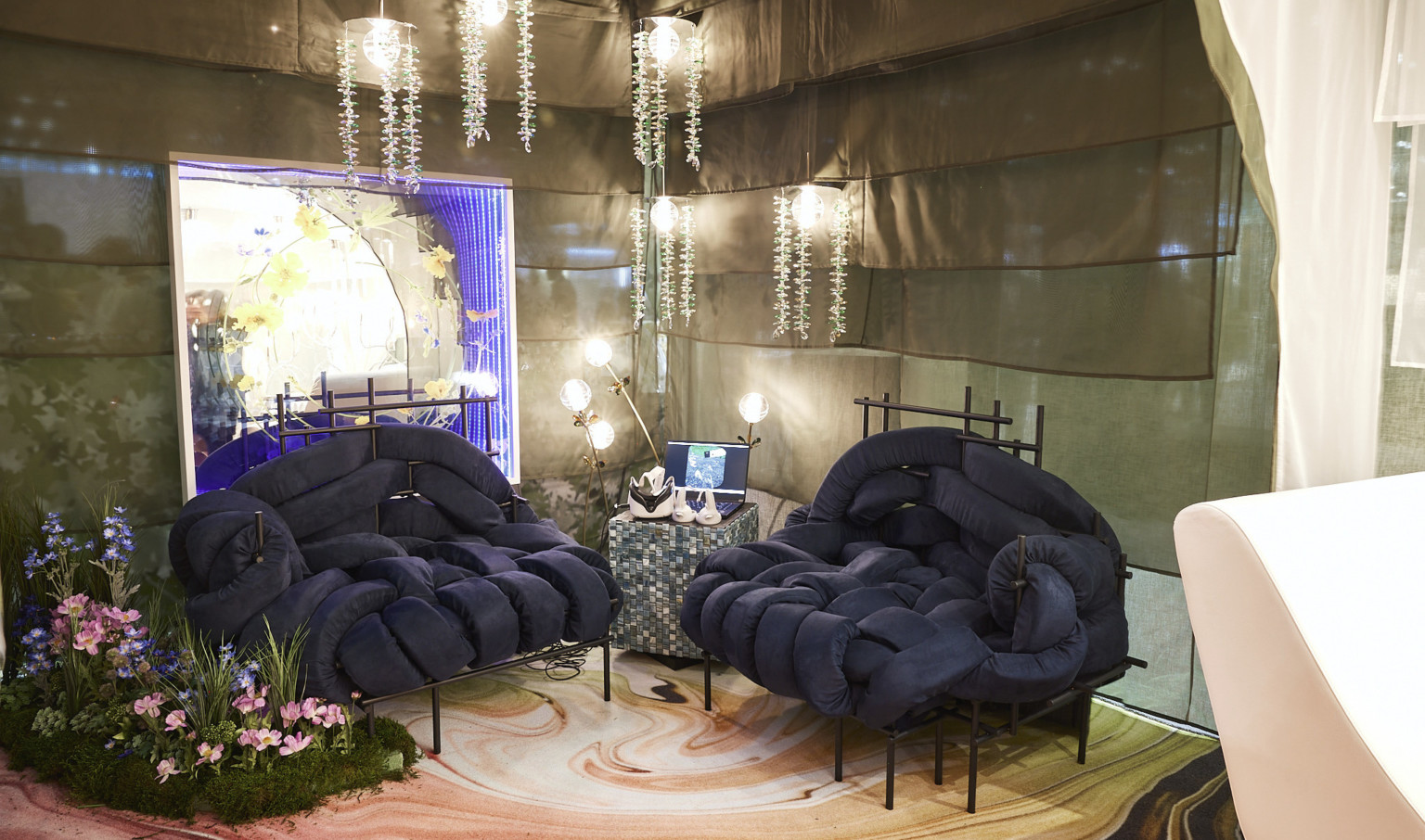 Navy woven cushioned chairs in corner of room with layered fabric partition walls. Greenery on floor, pocket wall accent, and hanging light