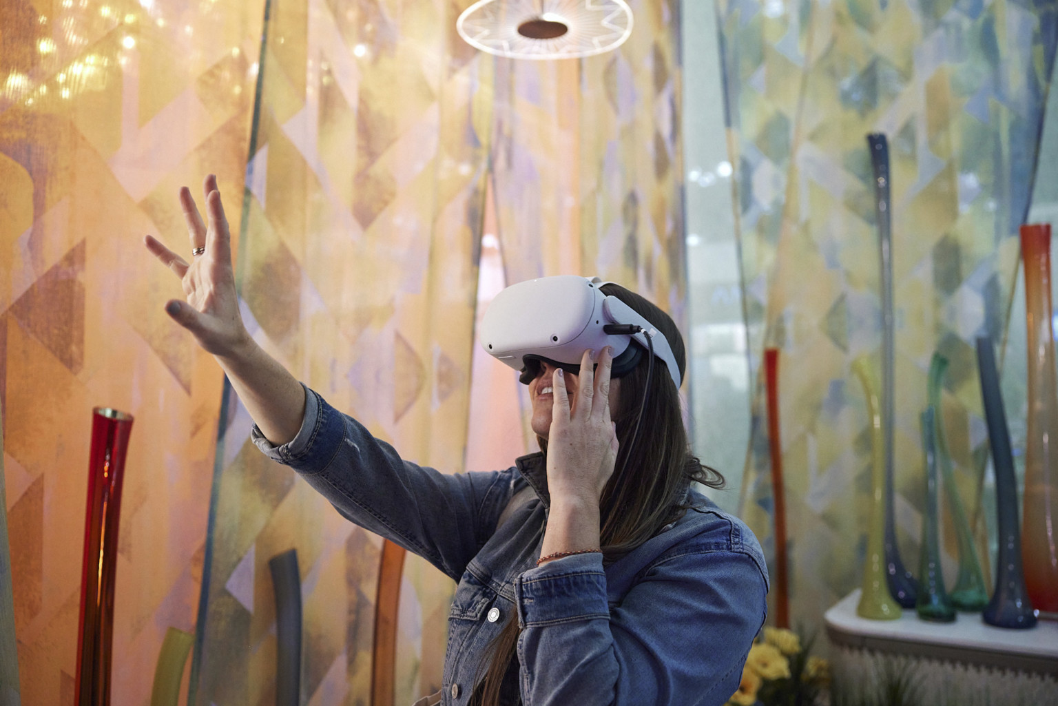 Woman using VR headset reaches out in room with geometric triangular patterned curtains in warm cool tone gradient. Organically shaped vases