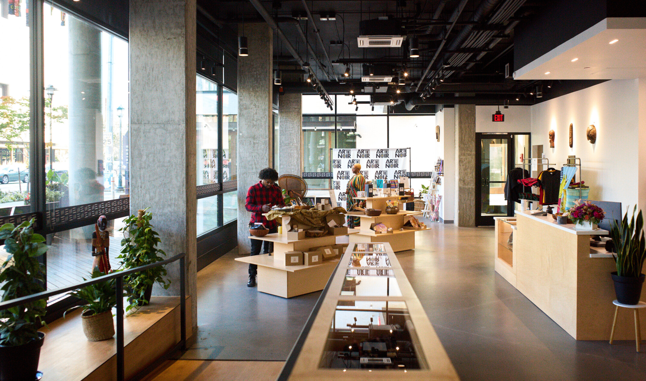 Store front in lobby of Arte Noir gallery. Blonde wood tiered tables and display cases in room with cement columns and large windows