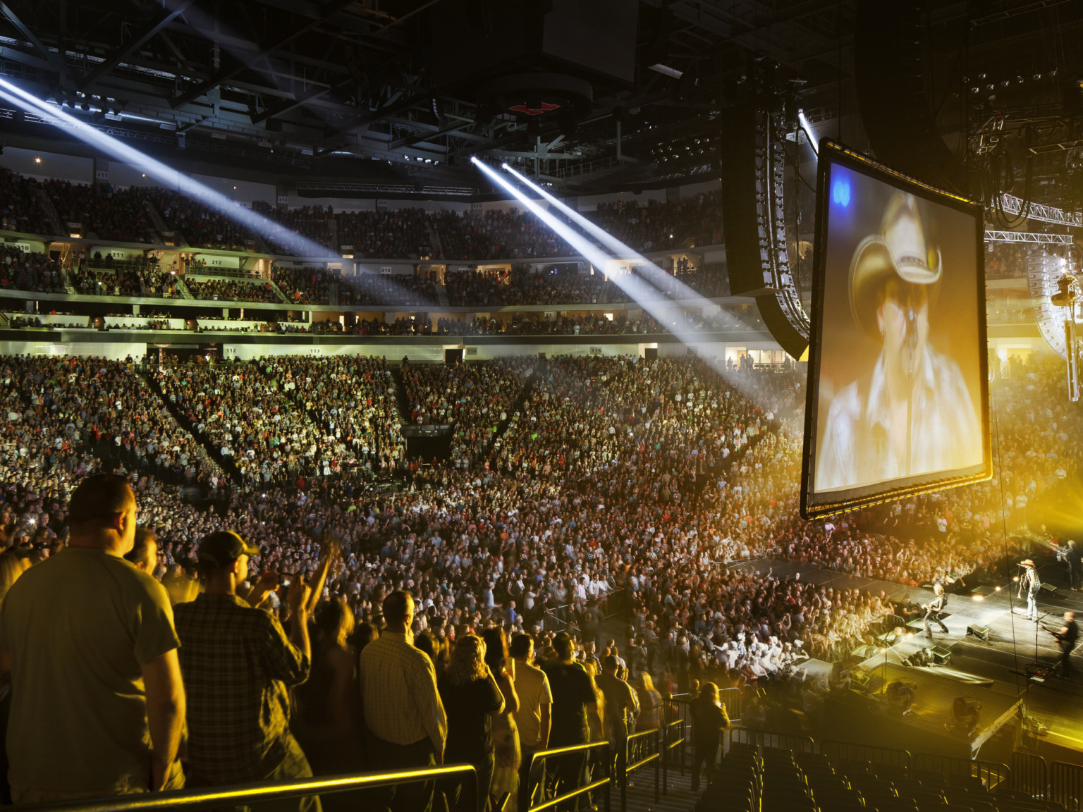 Interior, same space. Stadium seating filled during a concert performance on stage at center, lighting above and projection screen