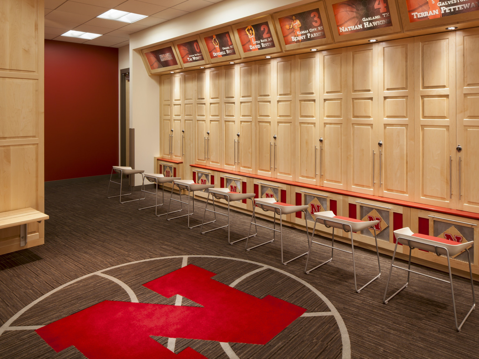 Locker room with blonde wood lockers each illuminated by recessed light, University of Nebraska logo at base and on carpet